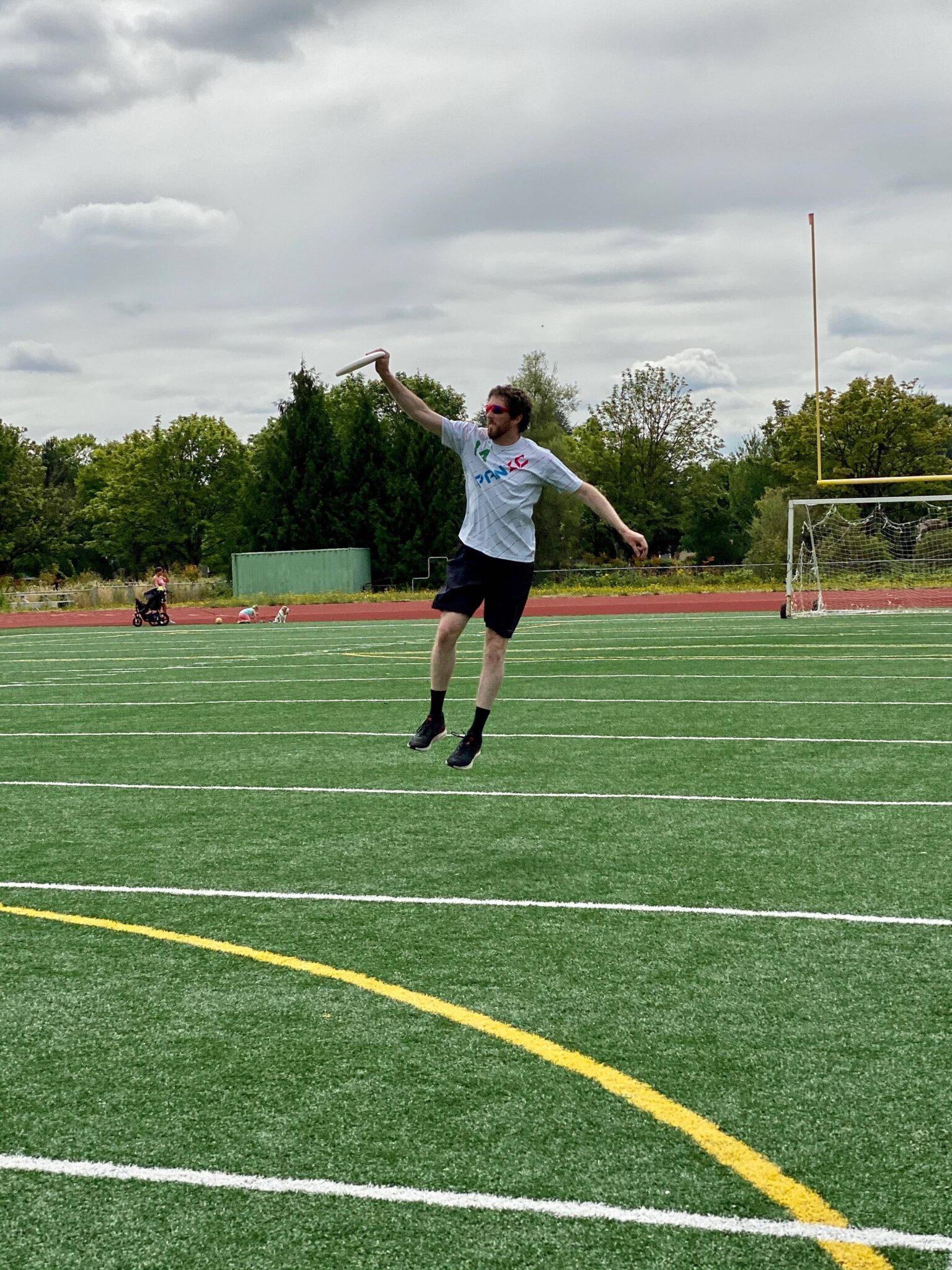 A photo of Will on a football field, jumping into midair as he plays ultimate frisbee. He wears a grey t-shirt, black shorts, black socks, and black sneakers.